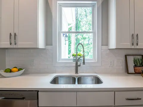 A white kitchen with a double steel sink under a window and a bowl of fruit on the counter