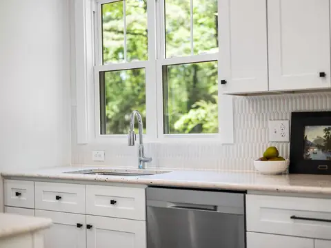 A white kitchen with the sink under a window onto green trees