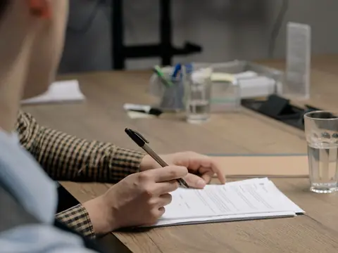 A hand with a pen going over a printed page at a wooden table beside a glass of water