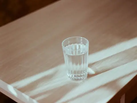 A glass of water casting a shadow on a sunlit wooden table