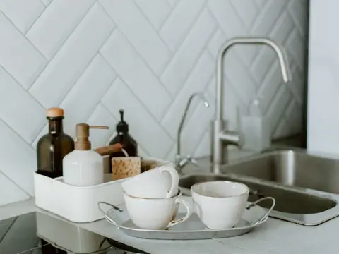 A steel faucet and sink beside tea cups and soap bottles against white tile